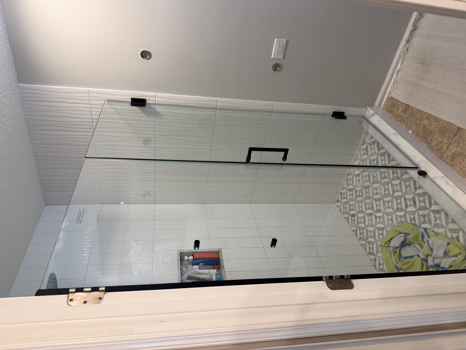 Overhead view of frameless shower glass panel with matte black hinges and hardware — installed over patterned tile floor